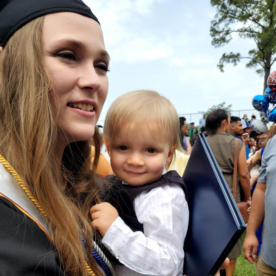 Tiffany Gillmore, wearing a graduation cap and gown, smiles while holding her young son.